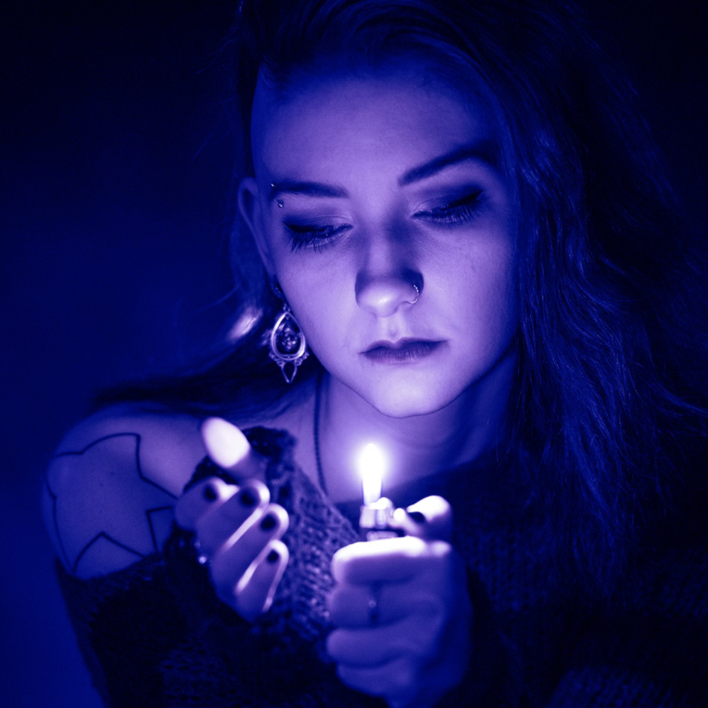 Catlea under blue lighting, holding a lit lighter and looking downward, with nose ring and ornate earring visible.