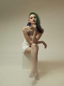 Catlea White Dress Green Hair Small - Bromsgrove Arts Catlea with green and blue hair sits on a white block, hand to chin, in a white dress, visible tattoos on shoulder and arm.