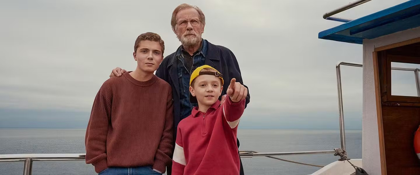 500 Miles - Bromsgrove Arts 500 miles - On a boat at sea, Bill Nighey as the Grandfather with grey beard & wearing a dark top standing behind his two grandsons, Roman Griffin Davis as Finn, wearing a brown jumper, and Dexter Sol Ansell as Charlie, wearing a red top and yellow baseball cap